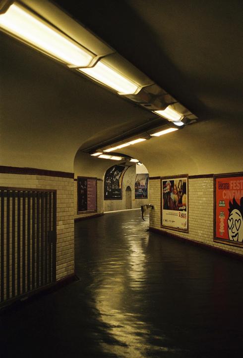 A long, curved underground tunnel is illuminated by fluorescent lights, with posters on the tiled walls and reflections on the wet floor.