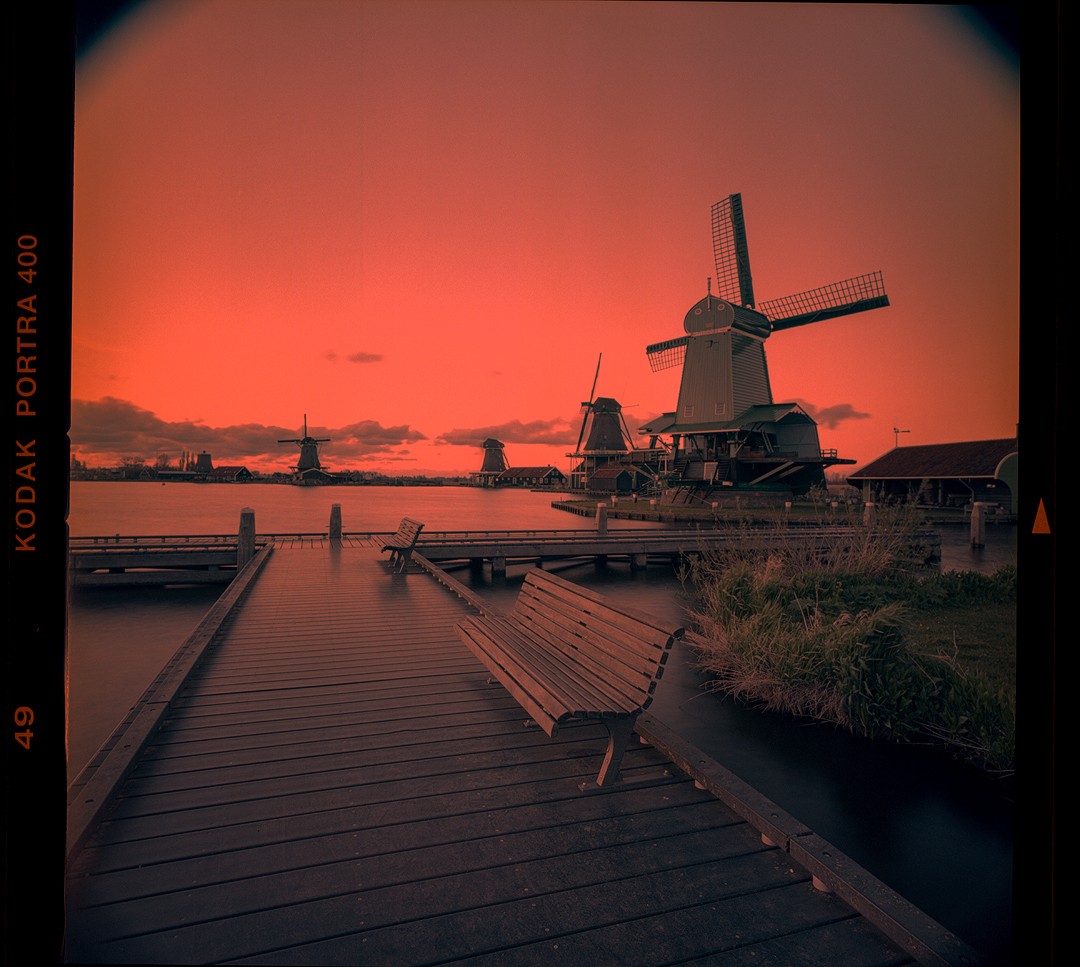 A wooden pier with benches extends into a body of water with several windmills under a vibrant orange sky.