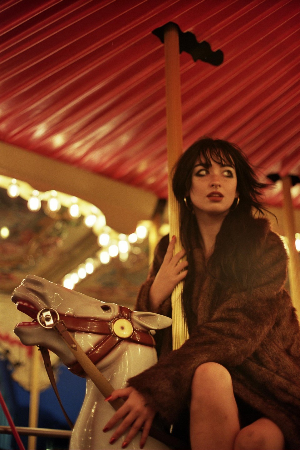 A woman with dark hair and a fur coat sits on a carousel horse at night.