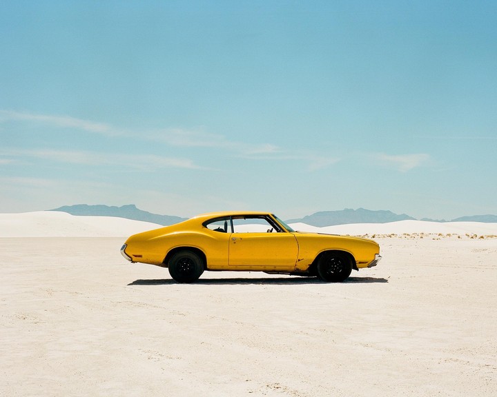 A yellow car is parked on a vast white sand desert with mountains in the background under a clear blue sky.