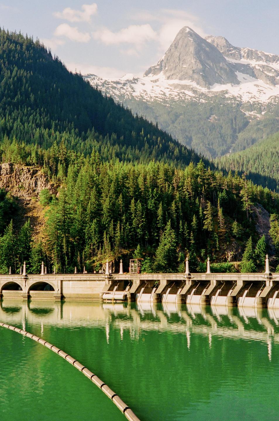 A dam with arches and pillars sits on a green lake, surrounded by a forest and a snow-capped mountain.