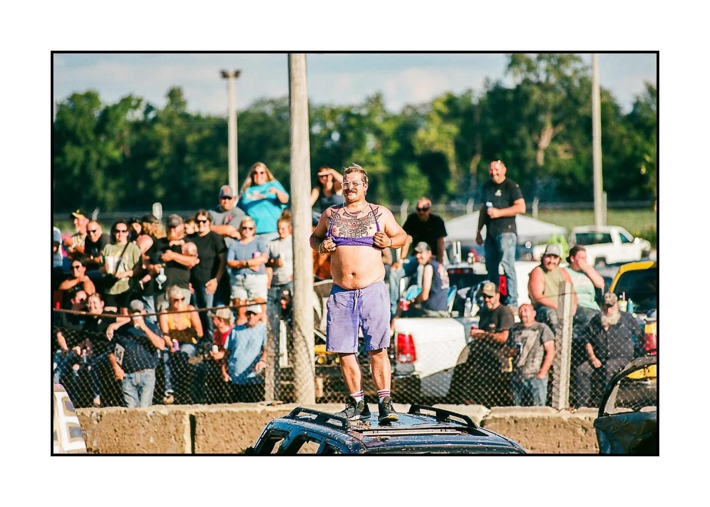 A man stands on the roof of a car in front of a crowd of people at a derby event.