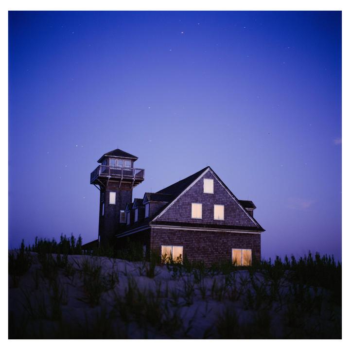 A house with a tower and illuminated windows stands on a sandy dune under a starry night sky.