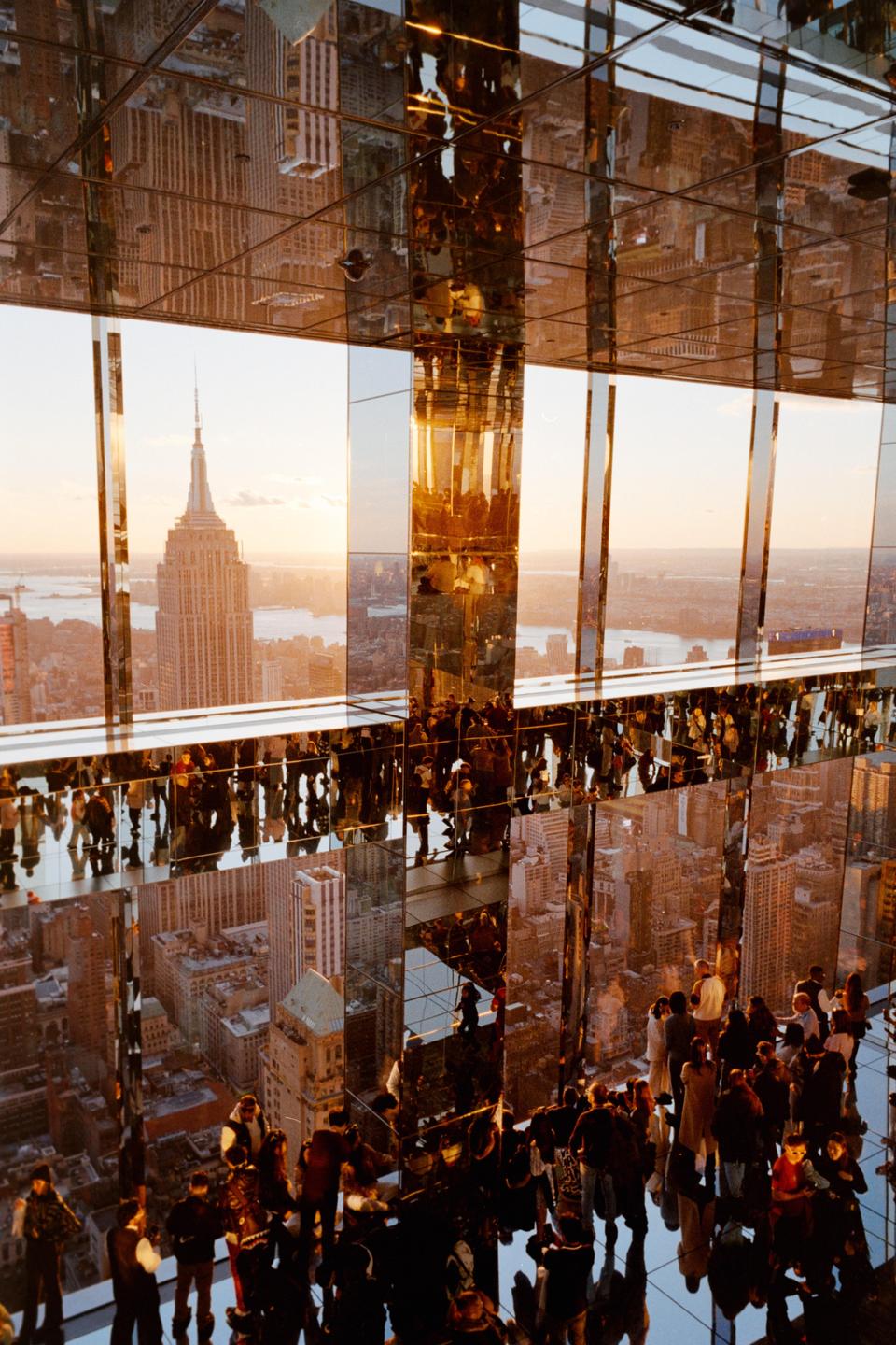 A crowd of people stands on a mirrored floor and looks out at the New York City skyline with the Empire State Building visible in the distance.