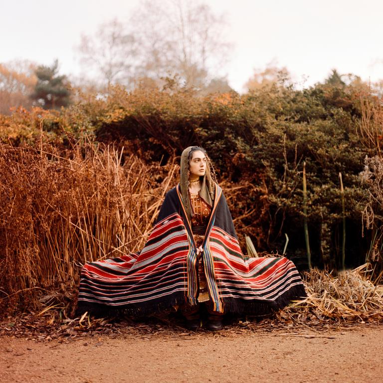 A woman wearing a colorful striped blanket and head covering sits on the ground surrounded by dry foliage.