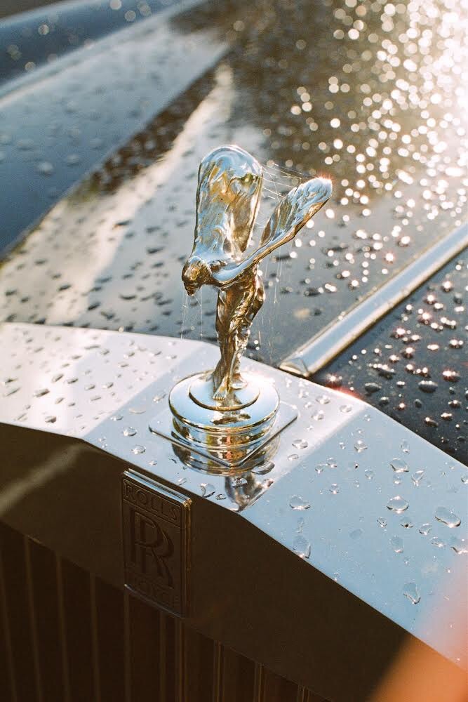 The Spirit of Ecstasy hood ornament on a Rolls-Royce is covered in water droplets and spiderwebs.