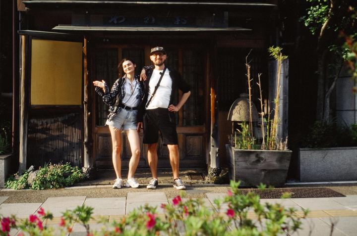 A couple stands in front of a traditional Japanese building with a stone lantern and bamboo plants to their right.