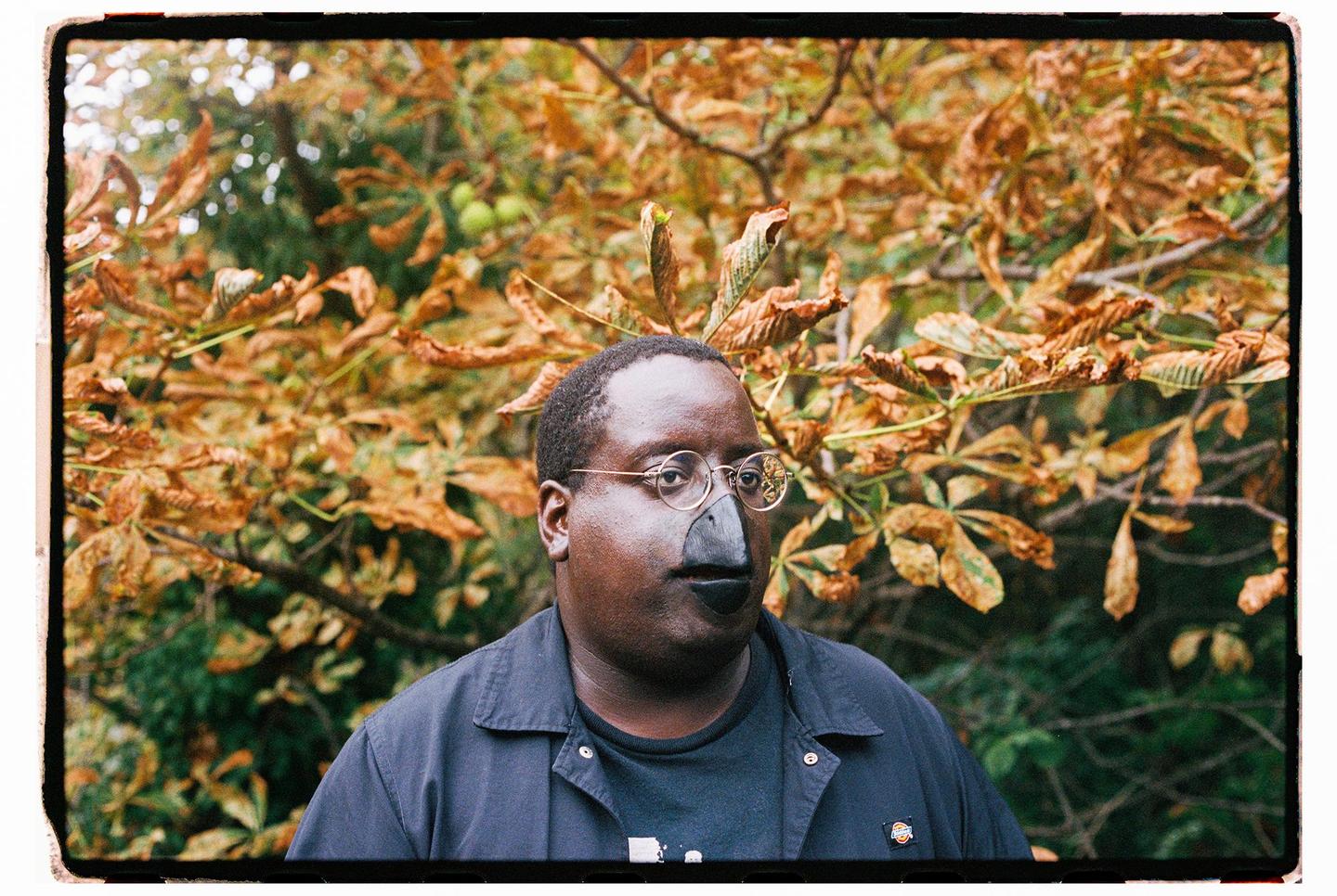 A man wearing glasses and a bird-like nose and mouth stands in front of autumn foliage.