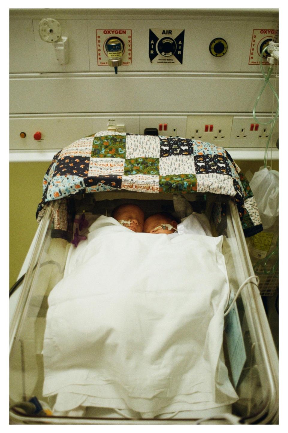 Two babies lie in a hospital incubator covered by a colorful quilt.