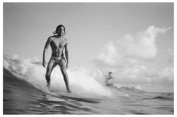 A man with long hair surfs a wave, with another surfer visible in the background.