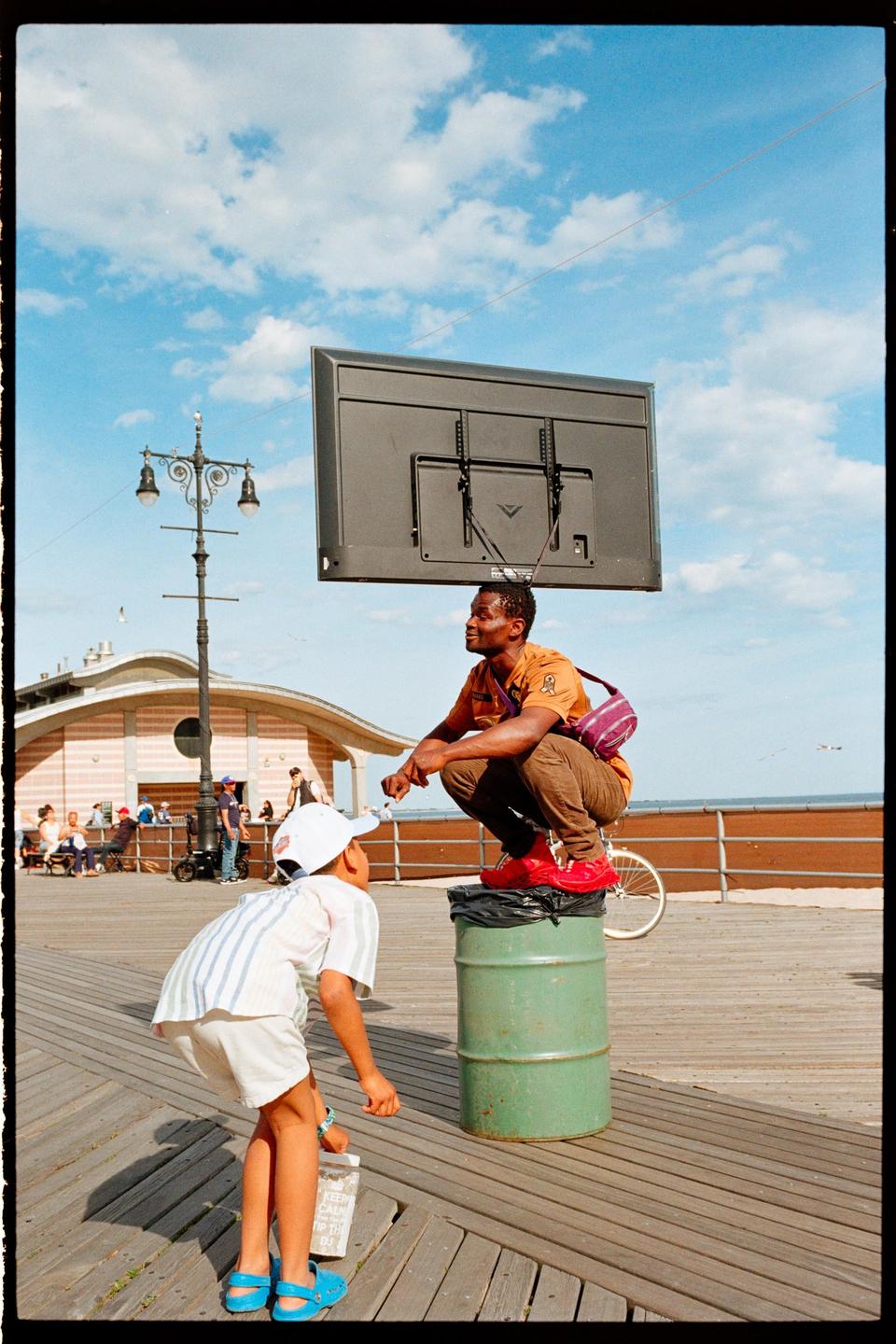 A man squats on a barrel with a television on his head while a boy looks up at him on a boardwalk.