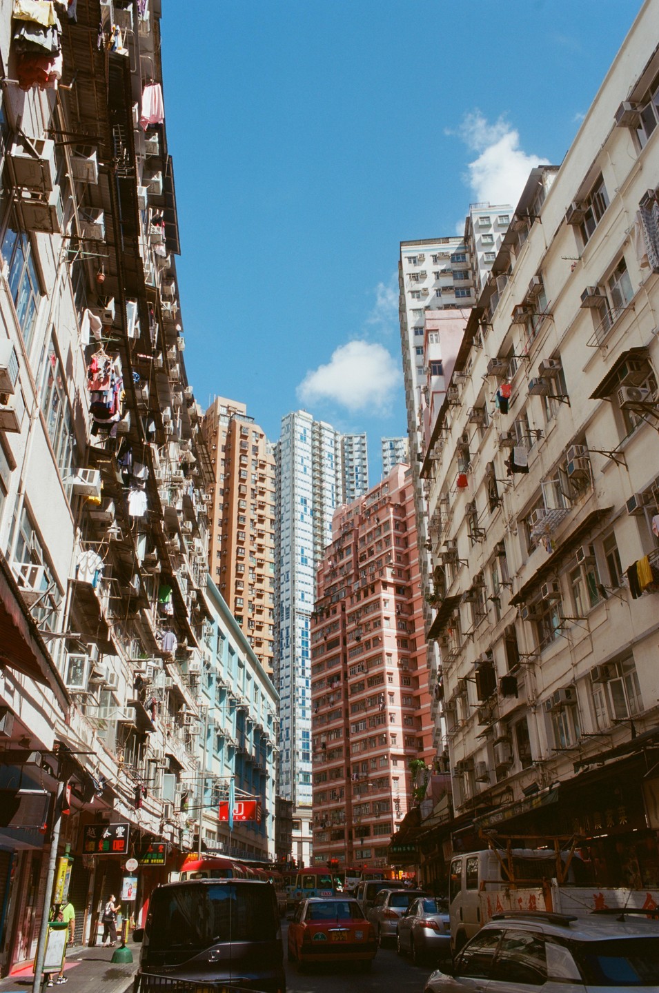 Tall buildings with clothes hanging from windows line a busy street with cars and pedestrians.
