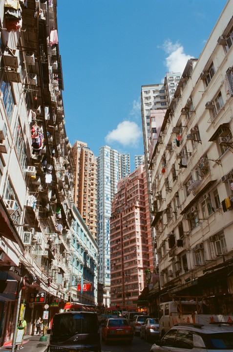 Tall buildings with clothes hanging from windows line a busy street with cars and pedestrians.
