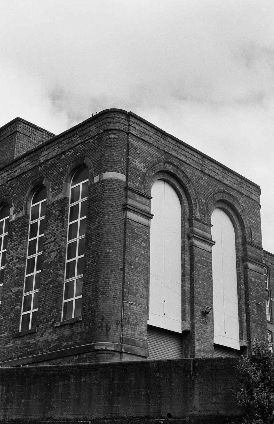 A large brick building with arched windows and a concrete wall in the foreground is shown from a low angle.