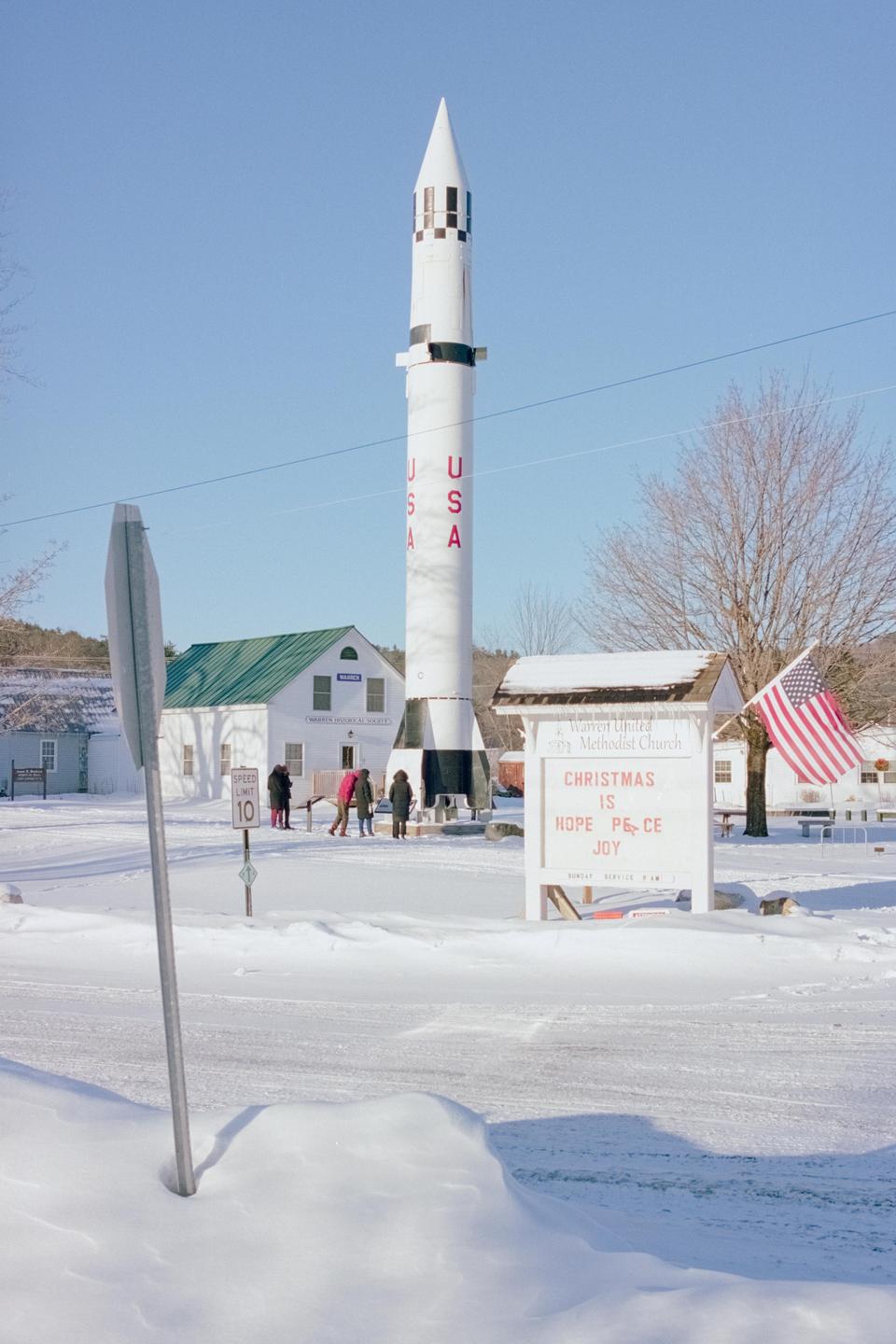A large white rocket stands in a snow-covered village with a church sign and an American flag.
