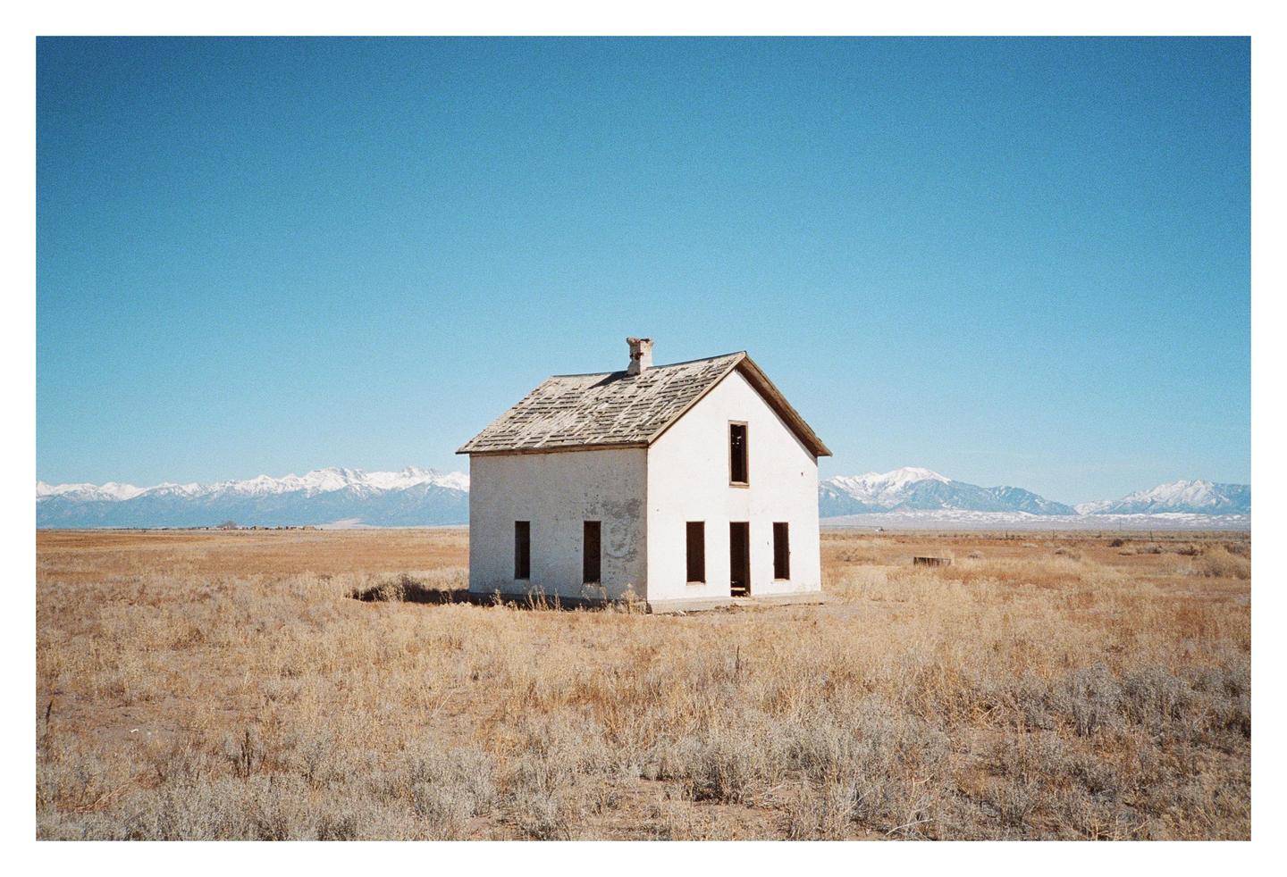A small, white, abandoned house stands alone in a dry, grassy field with snow-capped mountains in the distance under a clear blue sky.