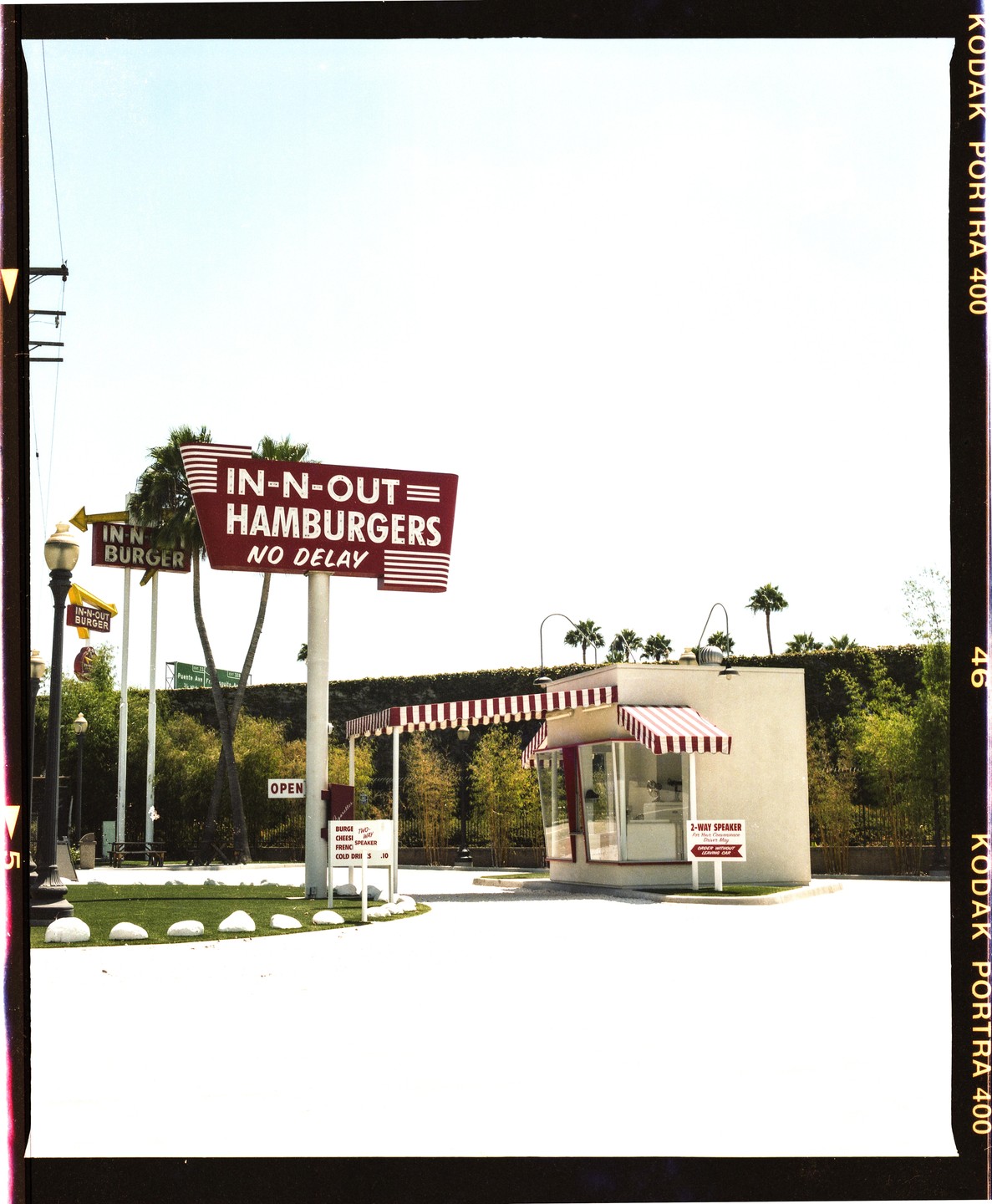 A drive-through hamburger stand with a large sign is visible under a bright sky.