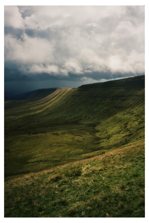 Sunlight illuminates a green mountain range under a cloudy sky.