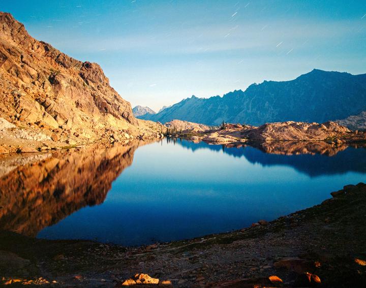 A long exposure shot of a lake at midnight shows star trails in the sky and mountains reflected in the water.