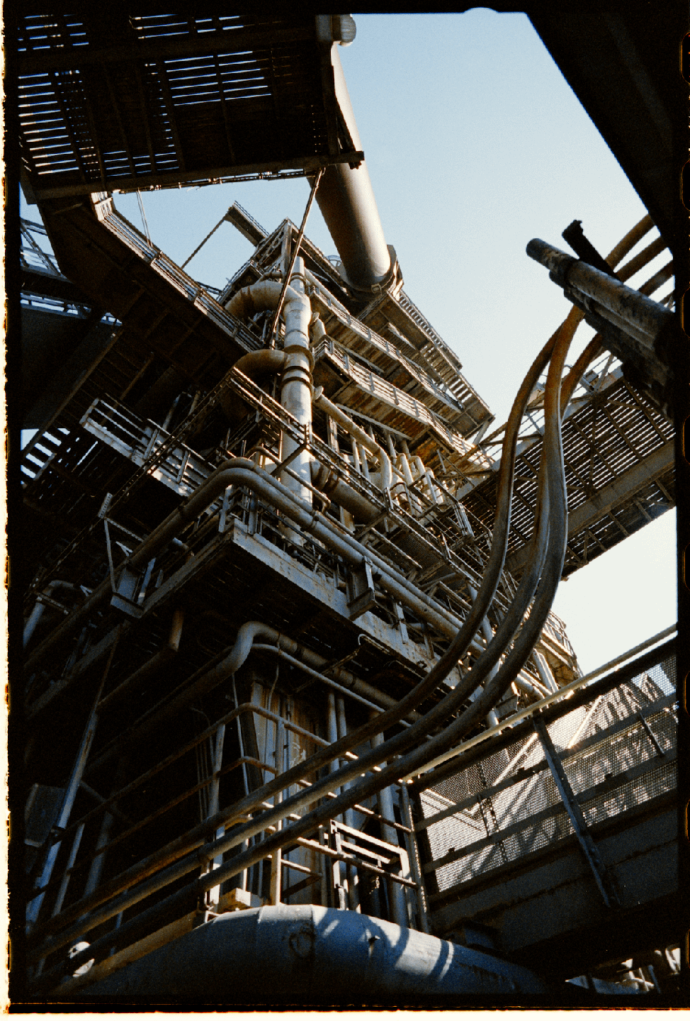 A low-angle view shows a complex industrial structure with many pipes and platforms against a clear sky.