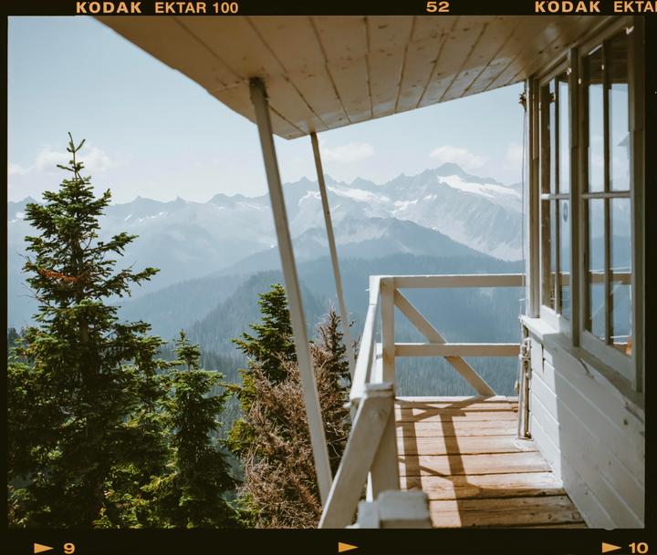 A wooden balcony of a building overlooks a mountain range with pine trees in the foreground.