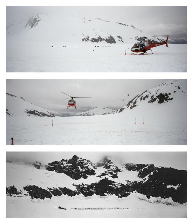 A red helicopter is parked on a snow-covered mountain with other helicopters flying in the distance.