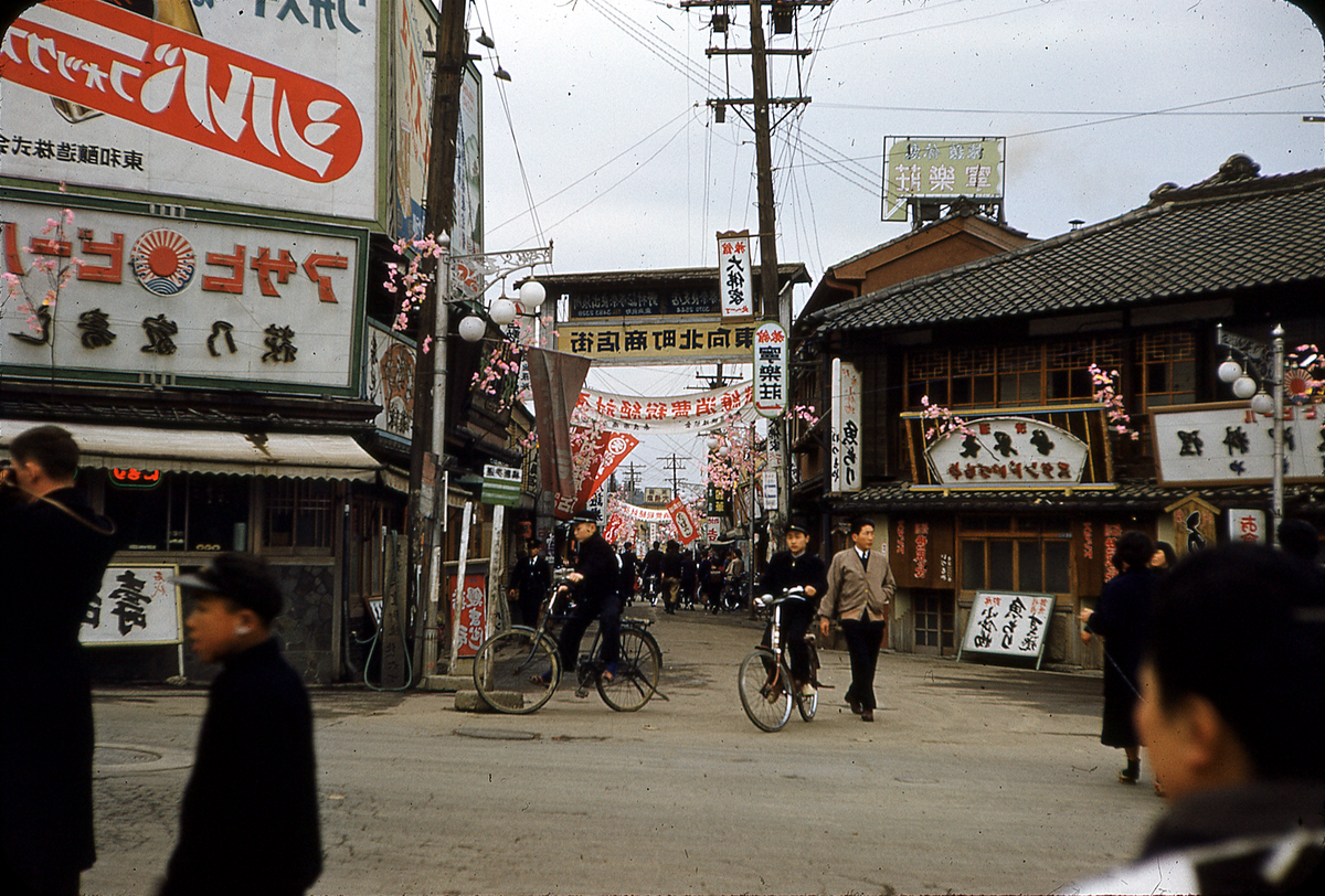 A street in Japan is bustling with people, bicycles, and shops adorned with banners and decorations.