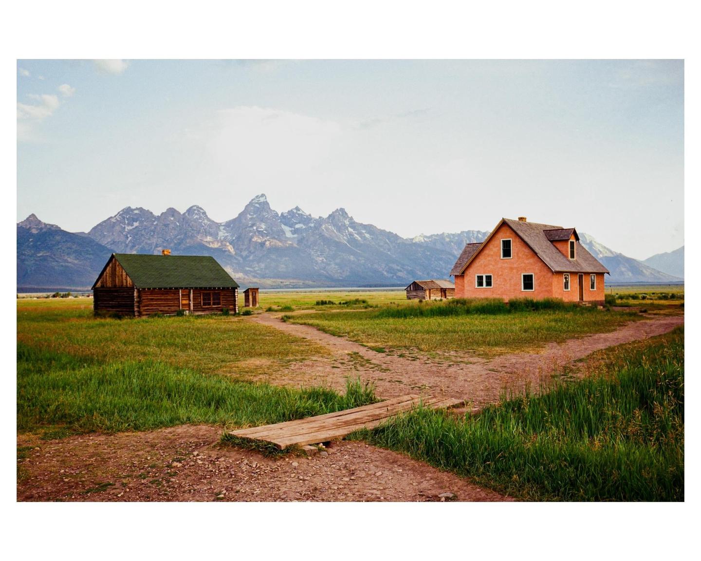 Two houses stand in a grassy field with a mountain range in the background.