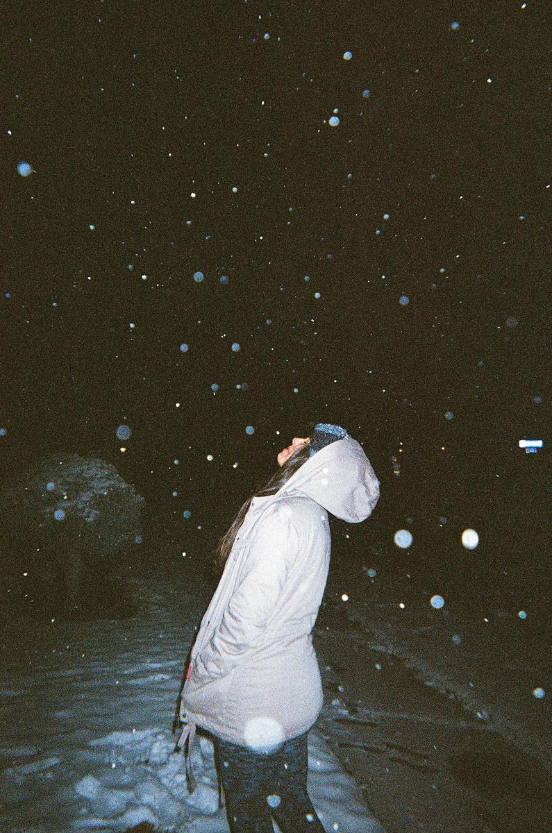 A woman in a hooded jacket stands in the snow at night, looking up at falling snowflakes.
