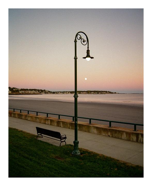 A street lamp and a bench overlook a calm body of water with a town in the distance under a pastel sky.