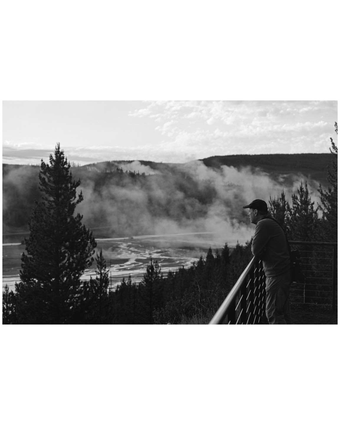 A man in a baseball cap leans on a railing overlooking a river valley with mist rising from the water and surrounding hills.