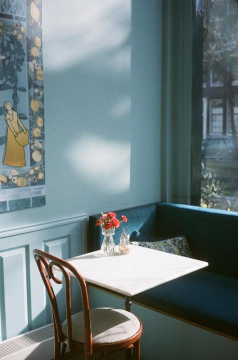 Sunlight streams into a cafe, illuminating a table with a vase of red flowers and a sugar dispenser, next to a blue banquette and a wooden chair.