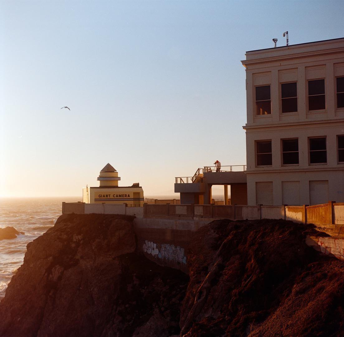 A building with a sign that reads "Giant Camera" stands on a cliff overlooking the ocean at sunset.