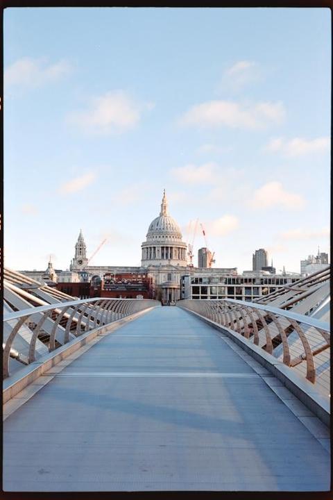 A long pedestrian bridge leads to a large domed building under a blue sky with scattered clouds.