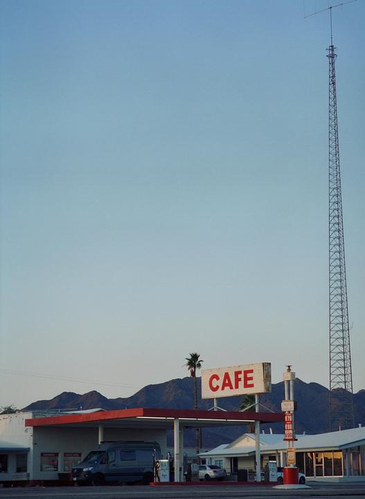 A gas station with a "CAFE" sign stands in front of mountains and a tall antenna.