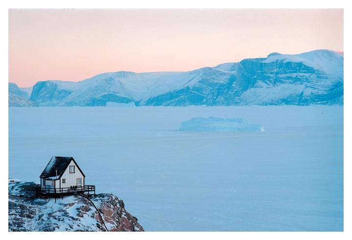 A small house sits on a snowy cliff overlooking a frozen body of water with mountains in the distance under a pink sky.