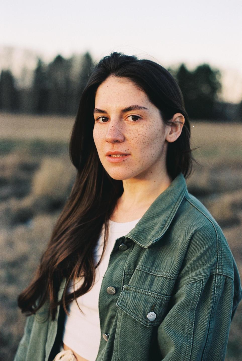 A woman with long dark hair and freckles wears a green jacket and white shirt while looking at the viewer.