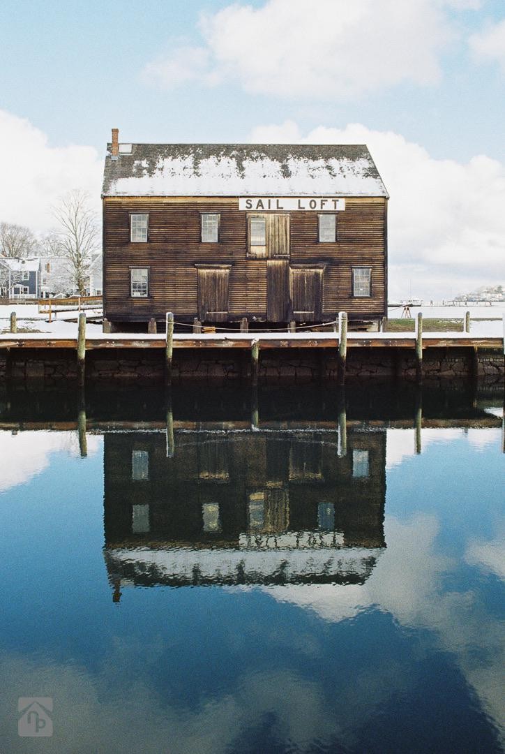 A wooden sail loft with a snow-covered roof is reflected in the calm water of a harbor.