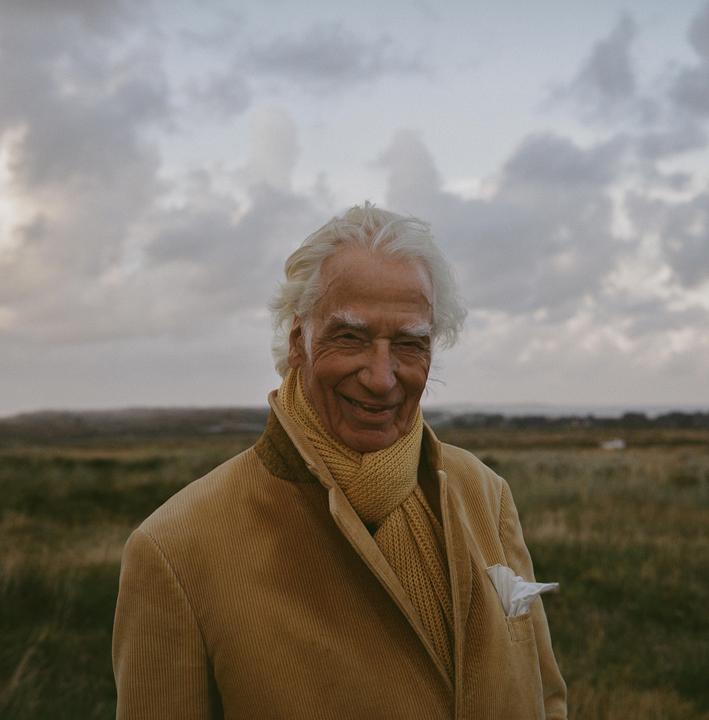 A smiling man with white hair and a yellow scarf stands in a field under a cloudy sky.