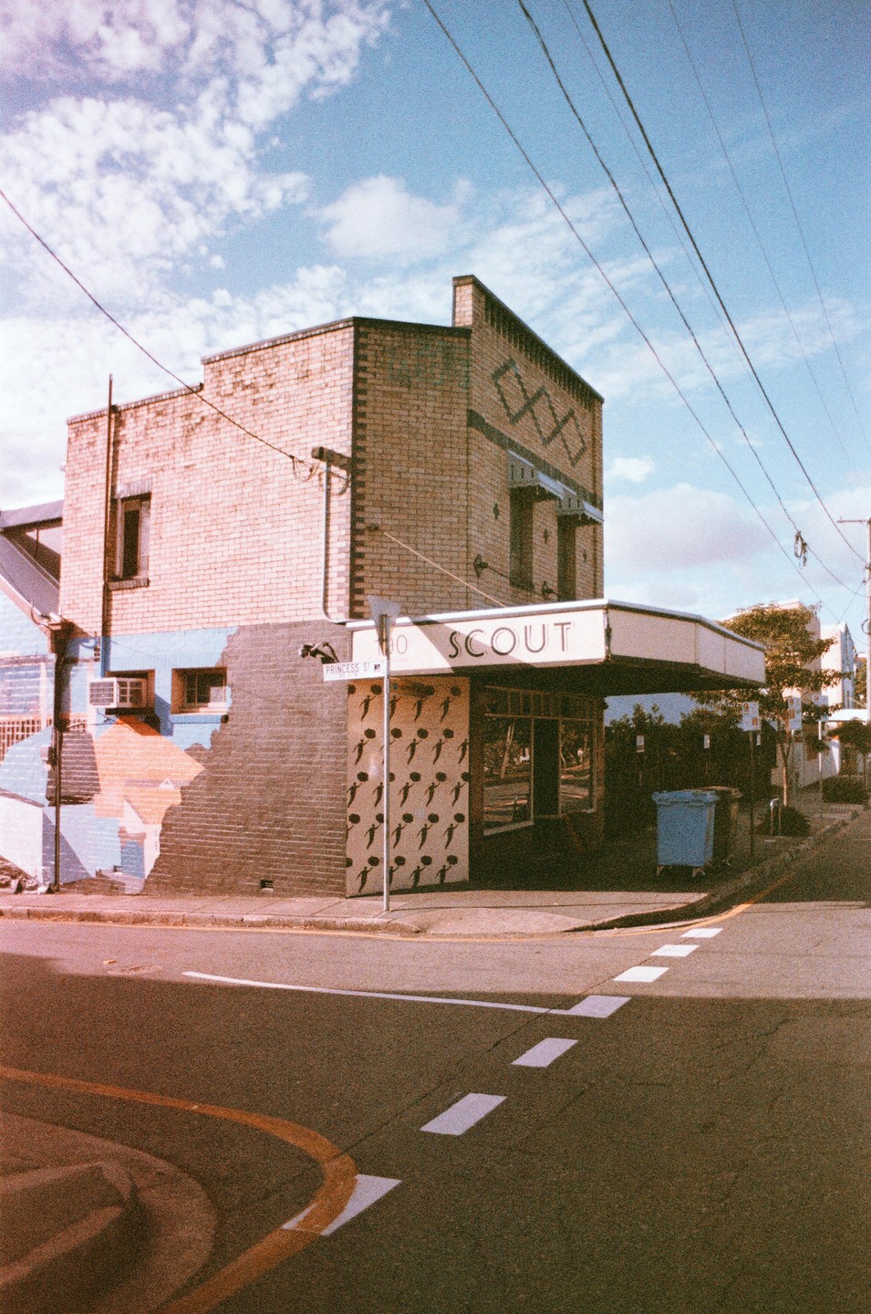 A brick building with a cafe on the ground floor stands at a street corner under a blue sky with clouds.