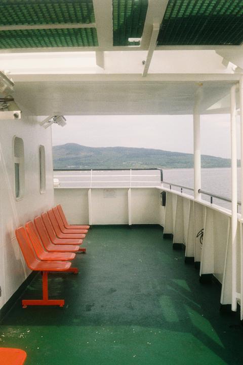 A row of orange seats sits on the deck of a ferry, with a body of water and mountains visible in the distance.