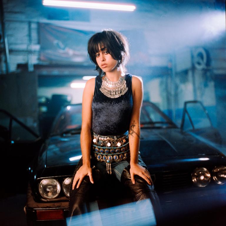 A woman with dark hair and a velvet top sits on the hood of a car in a garage.