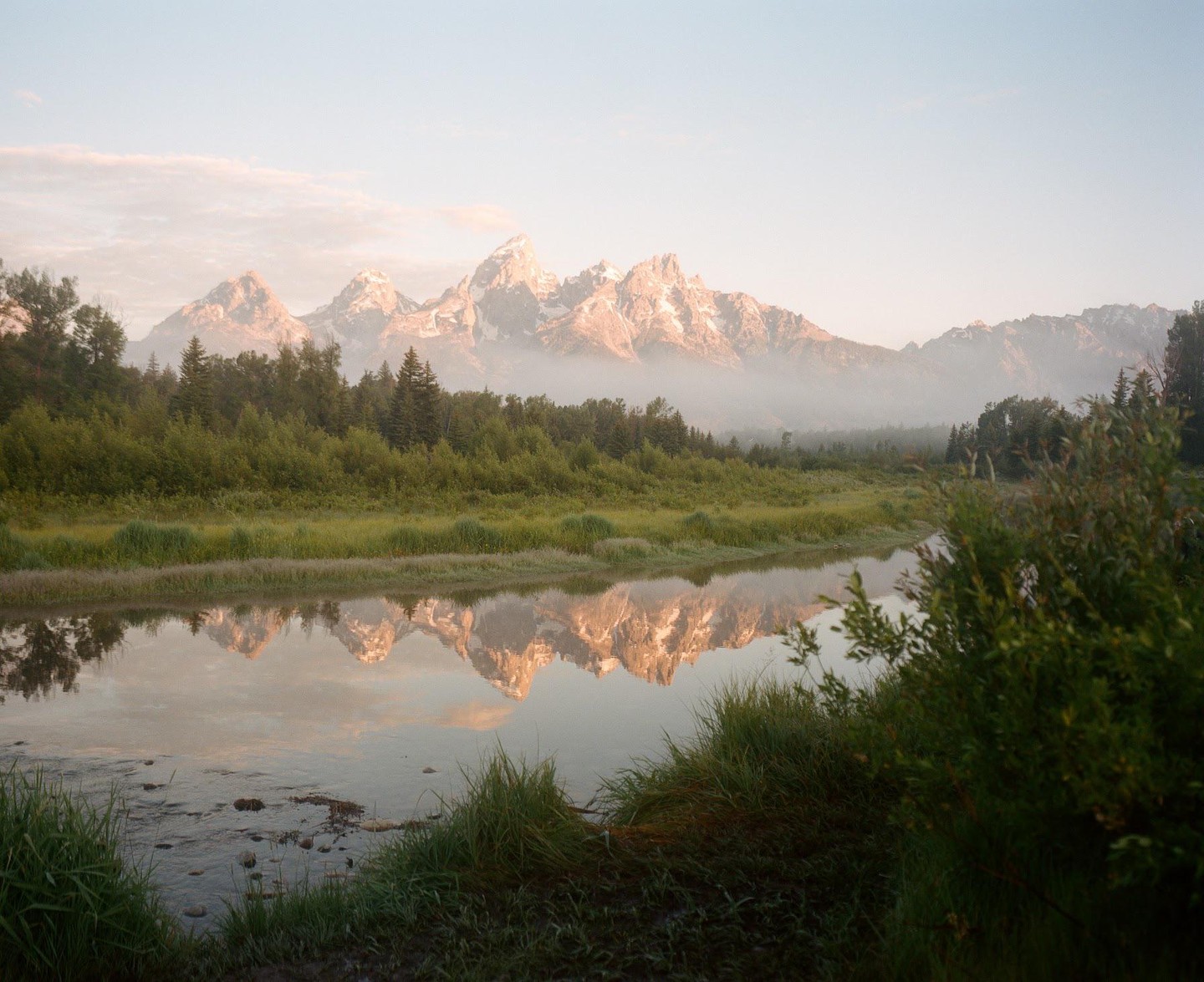 A mountain range with snow-capped peaks is reflected in a calm river surrounded by green trees and grass.