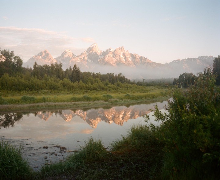 A mountain range with snow-capped peaks is reflected in a calm river surrounded by green trees and grass.
