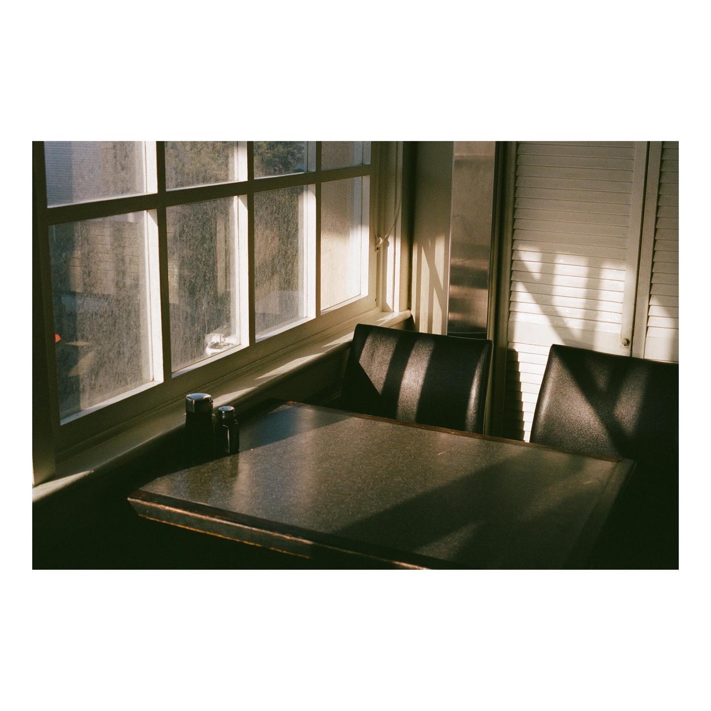 Sunlight streams through a window onto a diner table with two chairs and salt and pepper shakers.