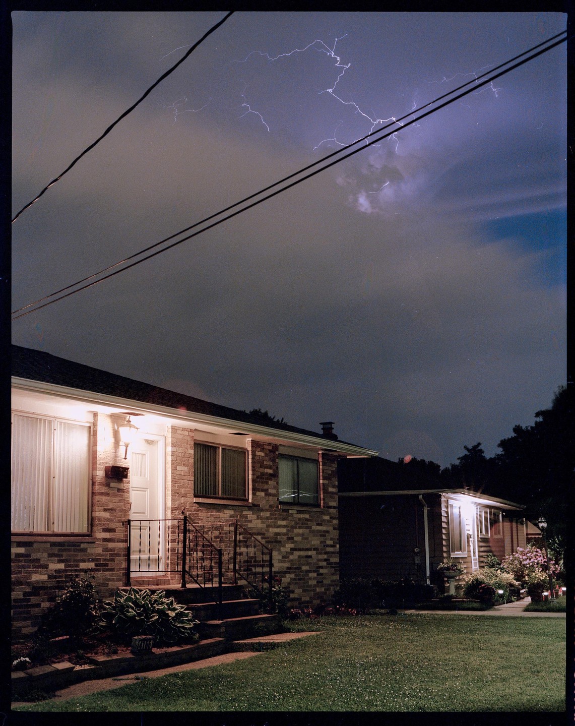 Lightning strikes across a dark, cloudy sky above two suburban houses at night.