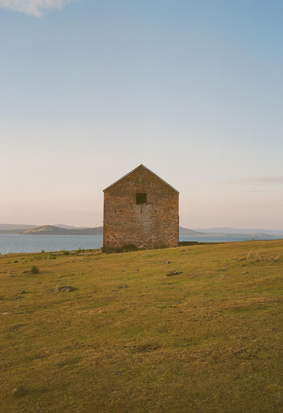 A brick house stands on a grassy hill overlooking a body of water with distant hills under a clear sky.