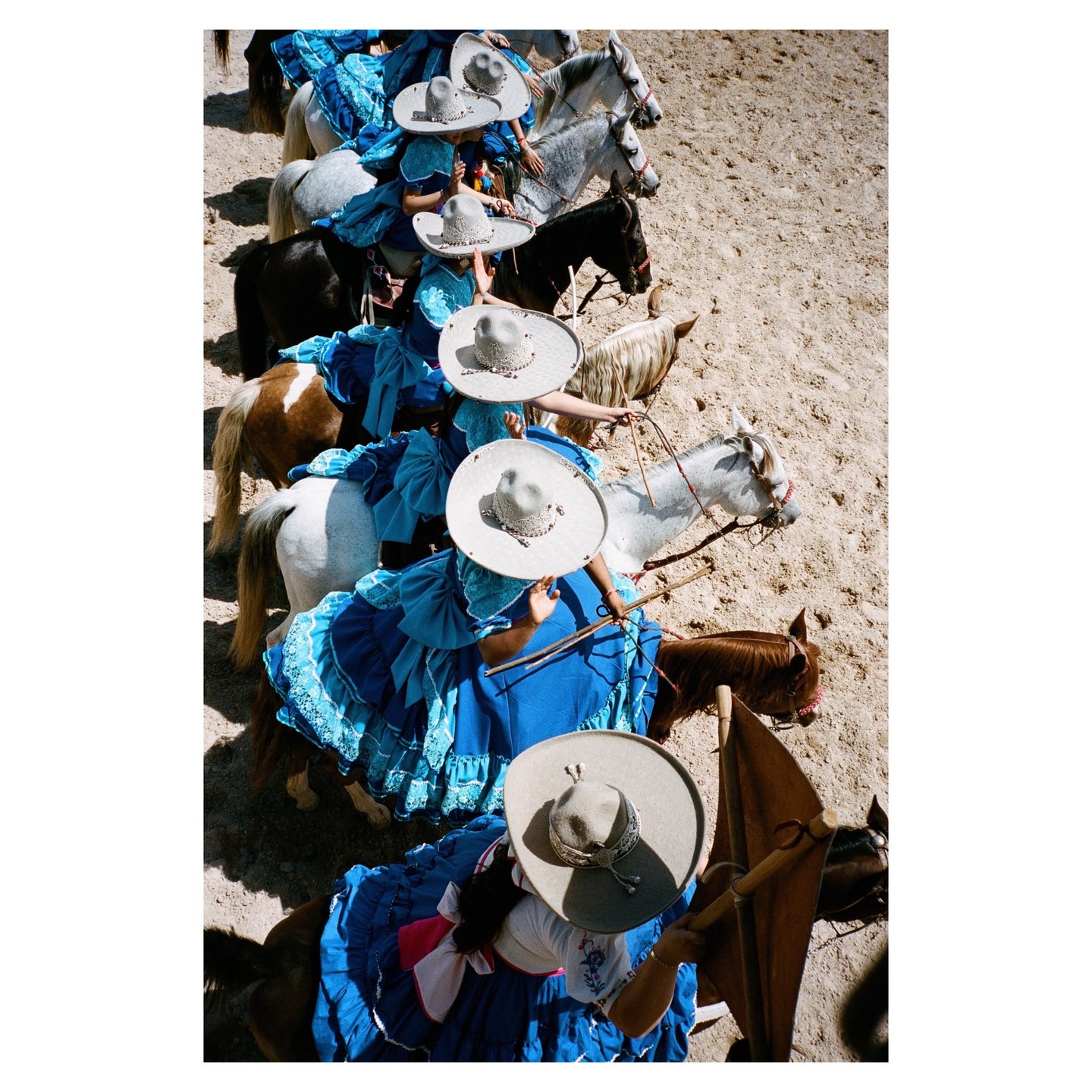 Several women in blue dresses and sombreros ride horses in a line.