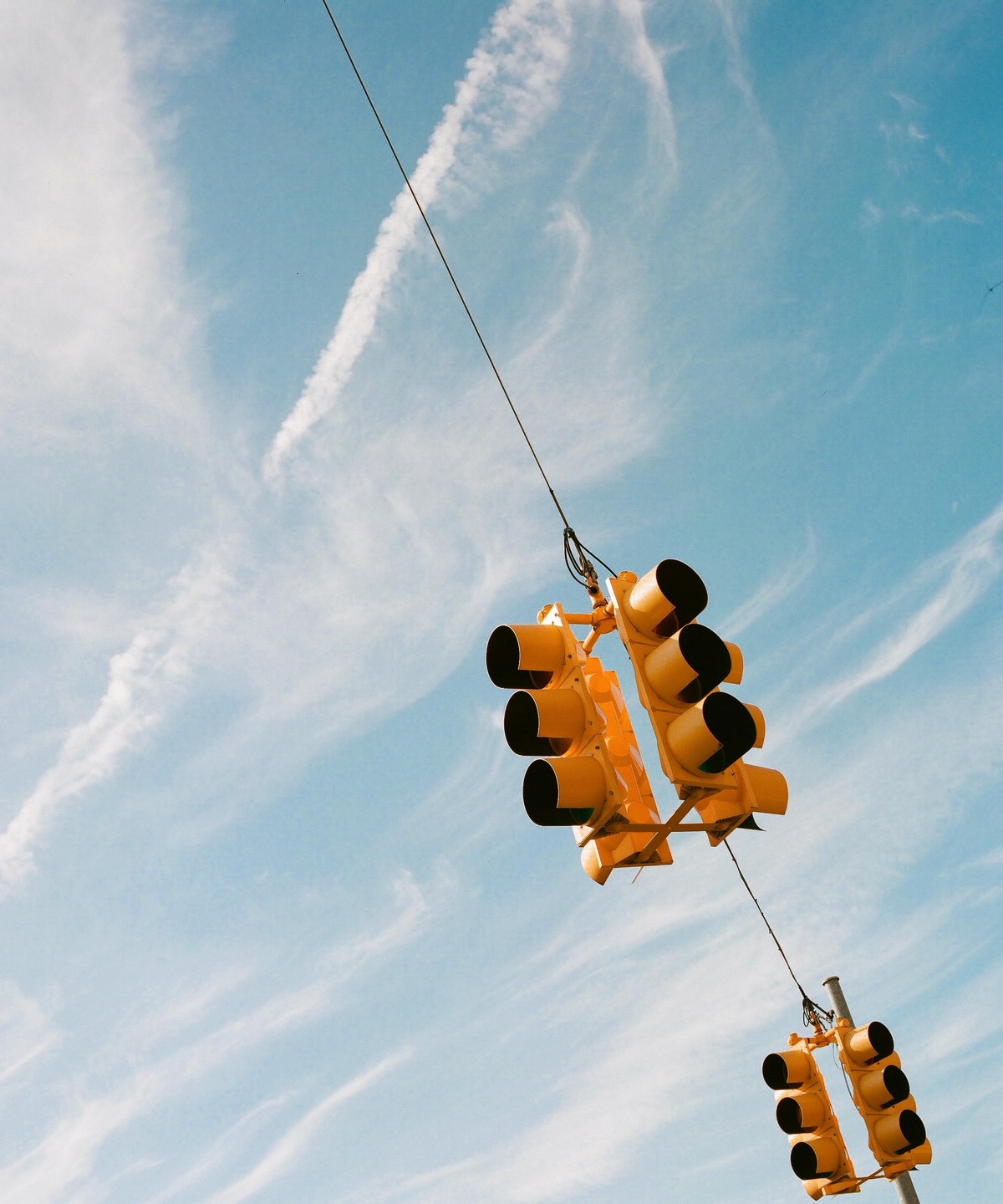 Two yellow traffic lights hang from wires against a blue sky with wispy clouds.