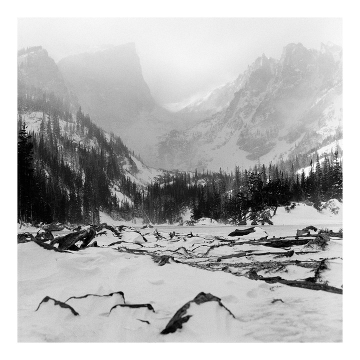 A snow-covered landscape features a frozen lake with logs and rocks in the foreground, surrounded by pine trees and mountains shrouded in mist.